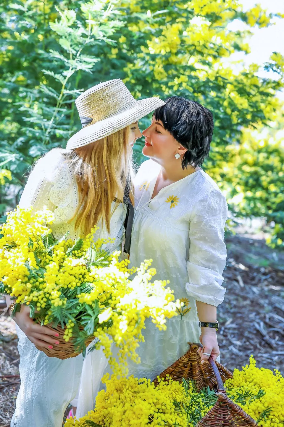 Two figures in white dresses gather yellow wattle flowers in a sunlit garden.