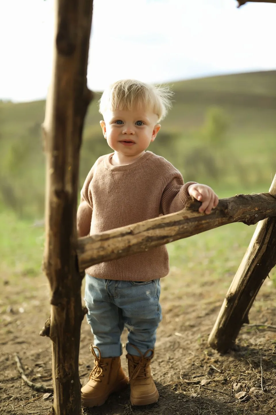 Toddler in brown sweater and jeans standing by wooden fence post in natural setting.