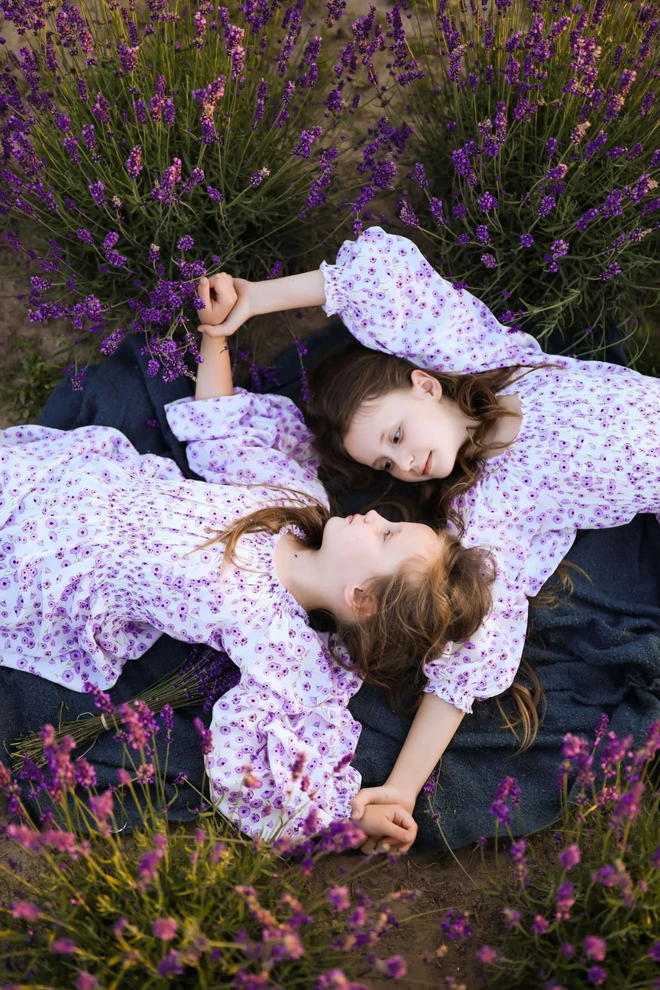 Two people in floral dresses rest head-to-head surrounded by vibrant purple lavender bushes.