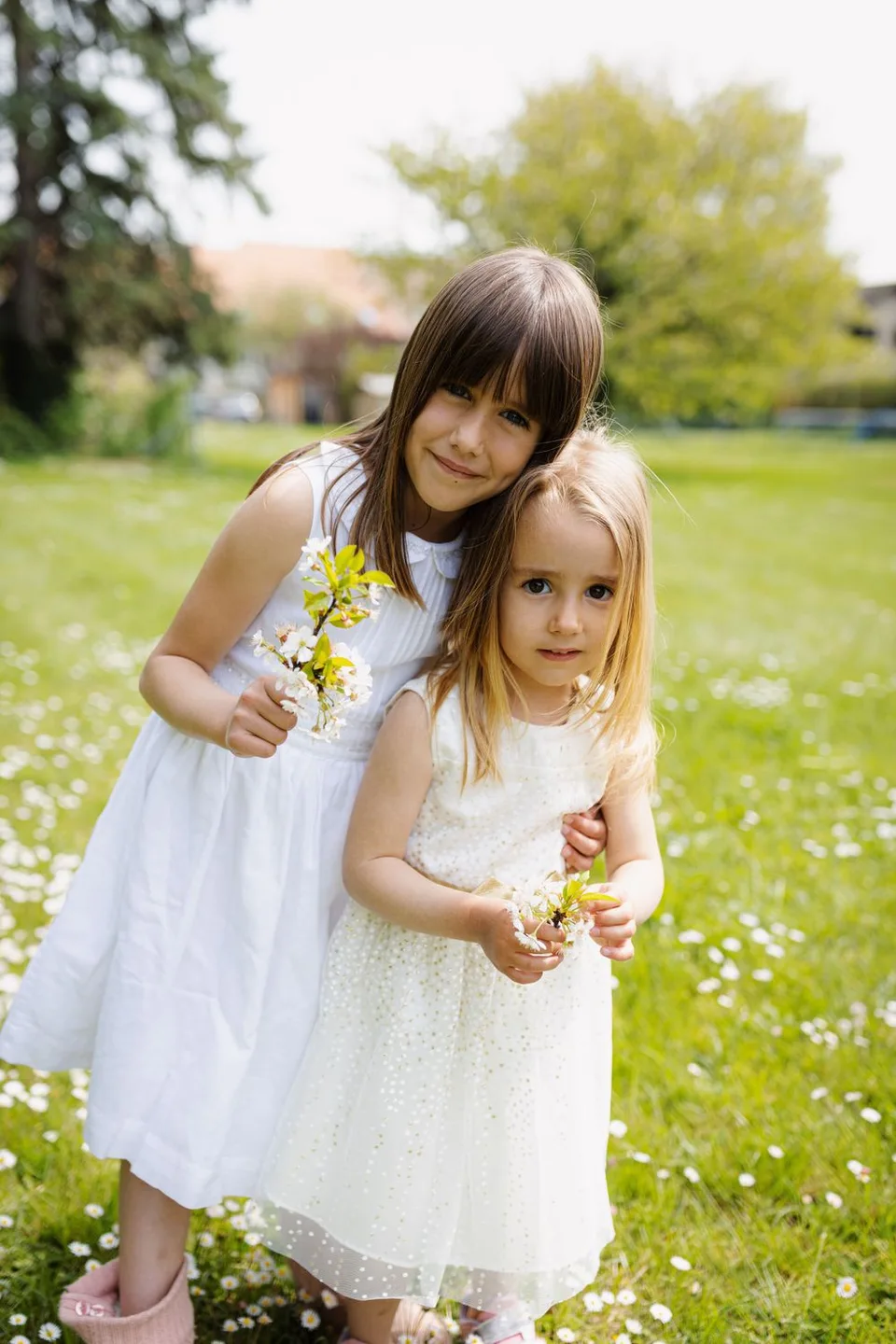 Two young friends in white dresses hold wildflowers while standing in a sunny meadow.