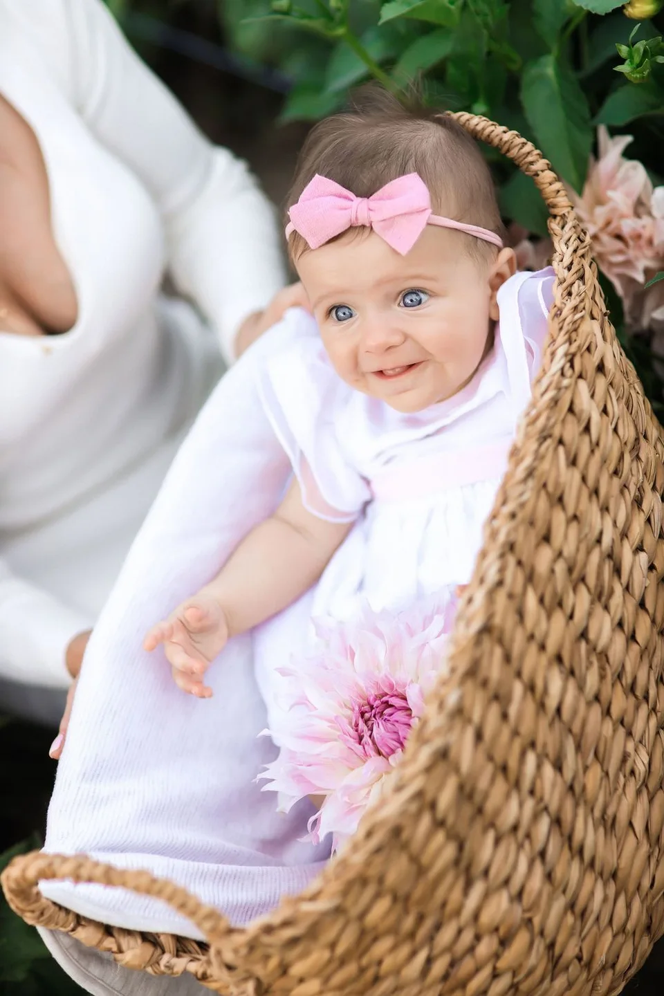 Baby in white dress with pink bow resting in woven basket with pink flower decoration.
