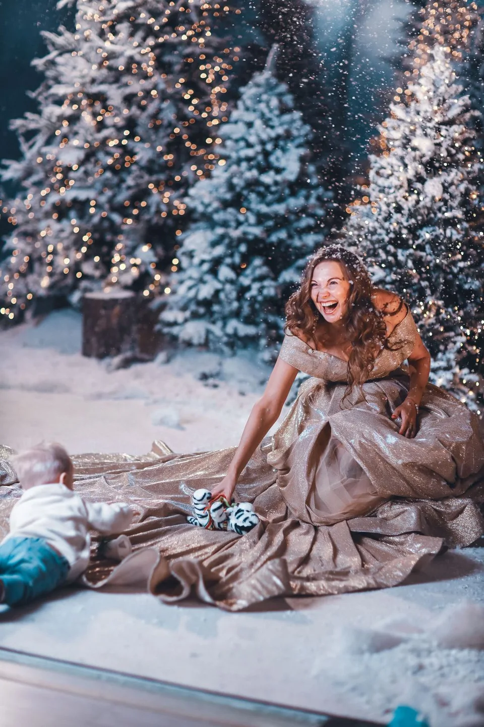 Person in elegant gown sitting among snow-covered Christmas trees with twinkling lights.