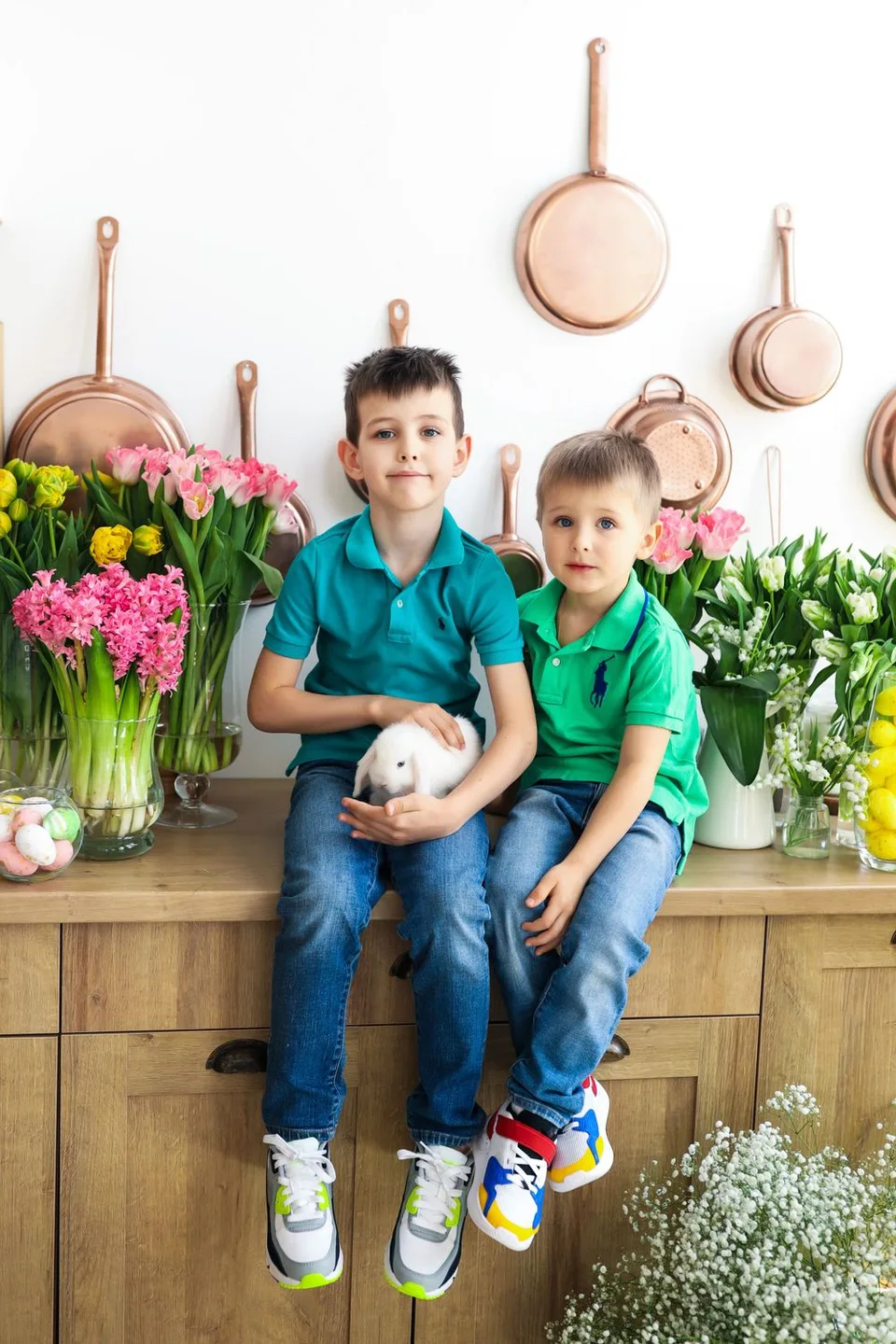Two siblings sitting on counter with bunny amid spring flowers and copper pans.