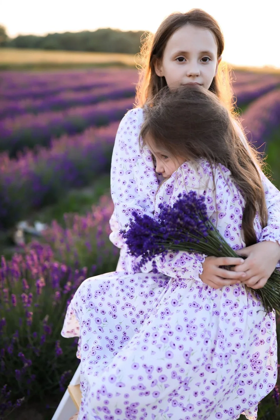Two people in floral dresses embrace while holding fresh-picked lavender in a purple field at sunset.