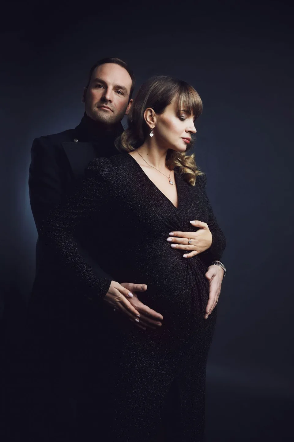 Dramatic maternity portrait in black formal wear against dark background.