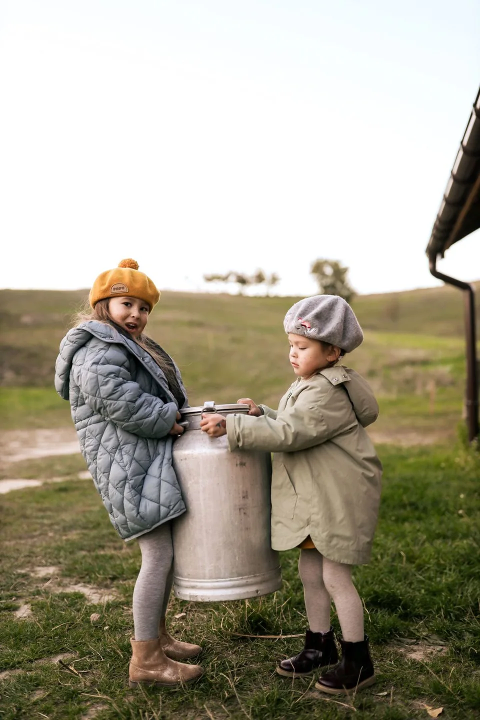 Two people in winter coats and beanies stand beside vintage milk can in countryside.