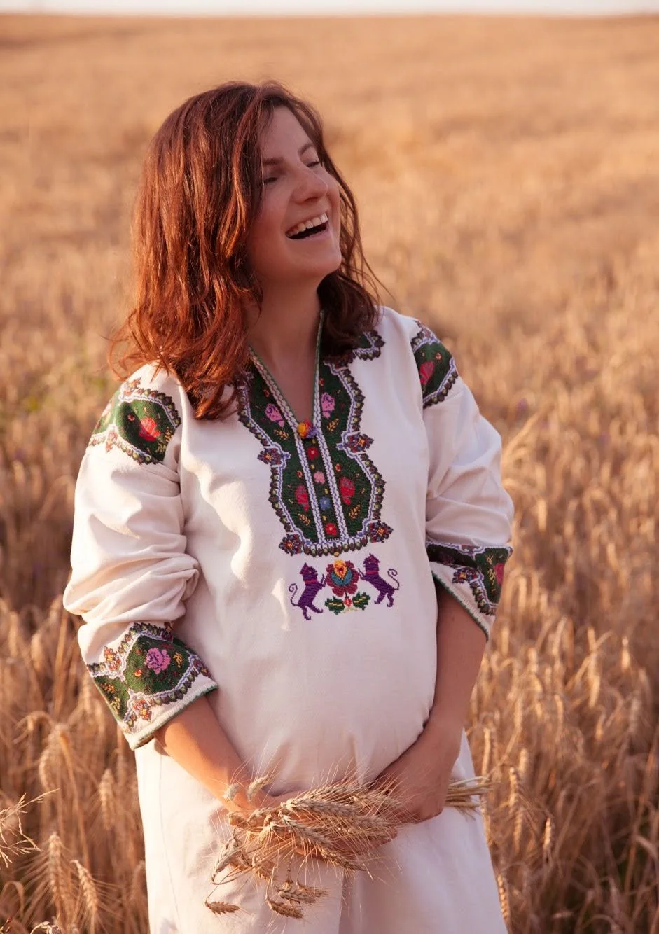 A pregnant woman in embroidered traditional dress holds wheat stalks in a golden field.