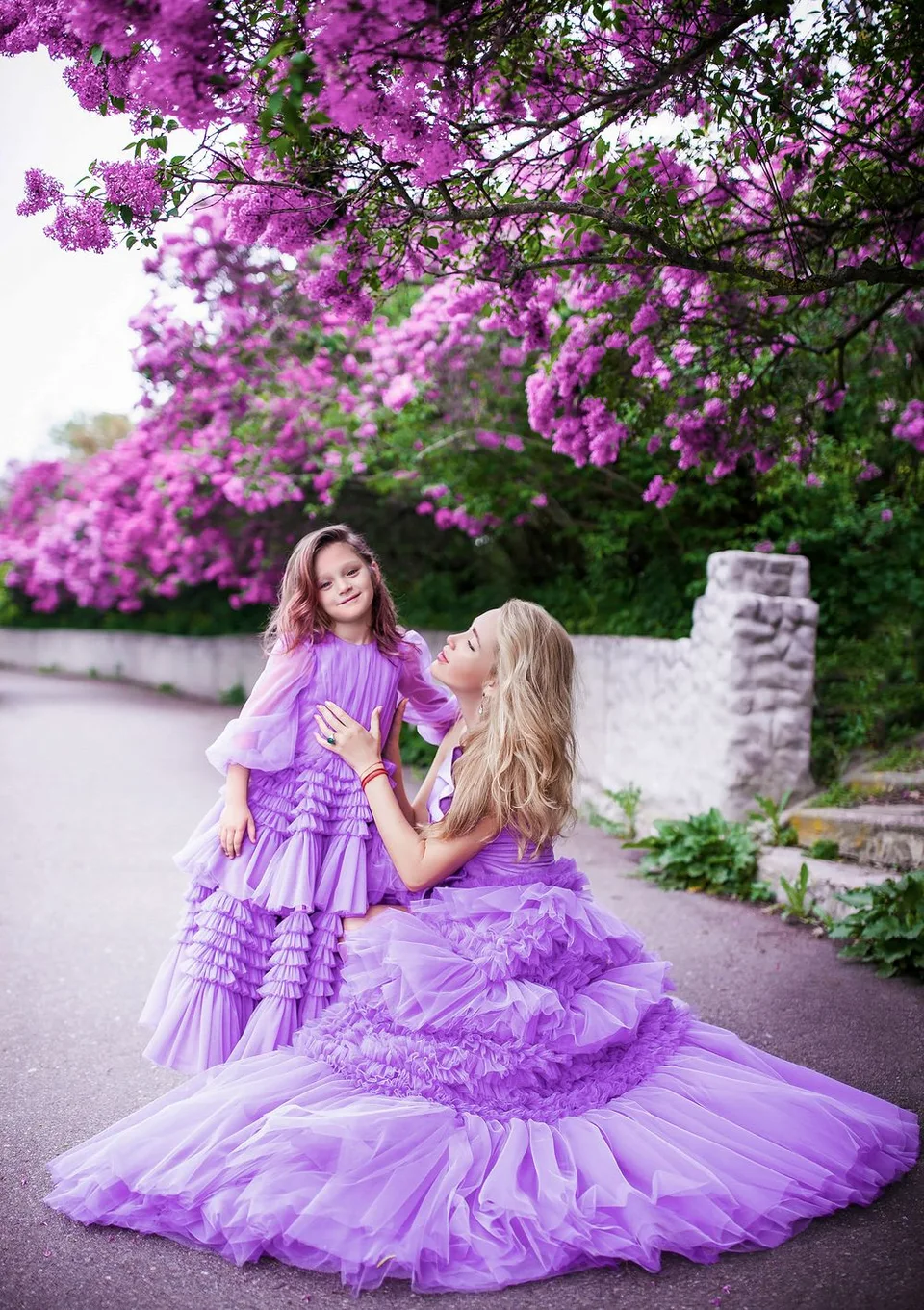 Two people wearing matching purple ruffled gowns pose beneath vibrant purple bougainvillea flowers.