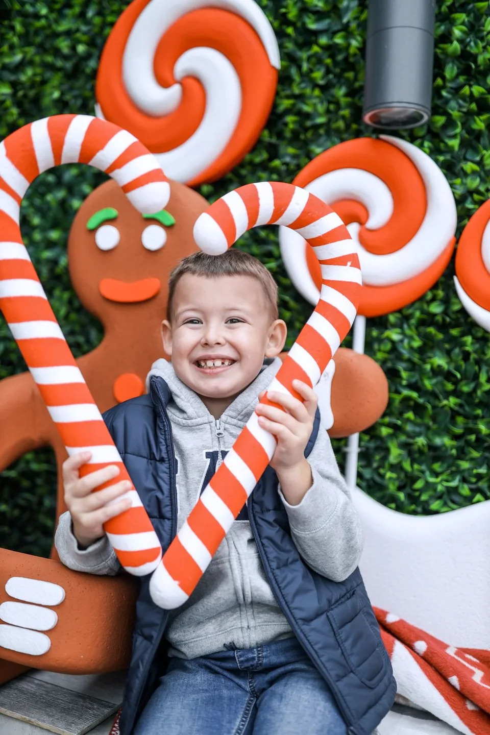Child holds large candy canes against decorative gingerbread man holiday display.