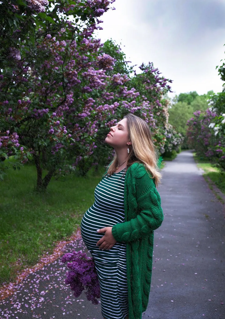 Expectant mother in striped dress and green cardigan poses by blooming lilac trees.
