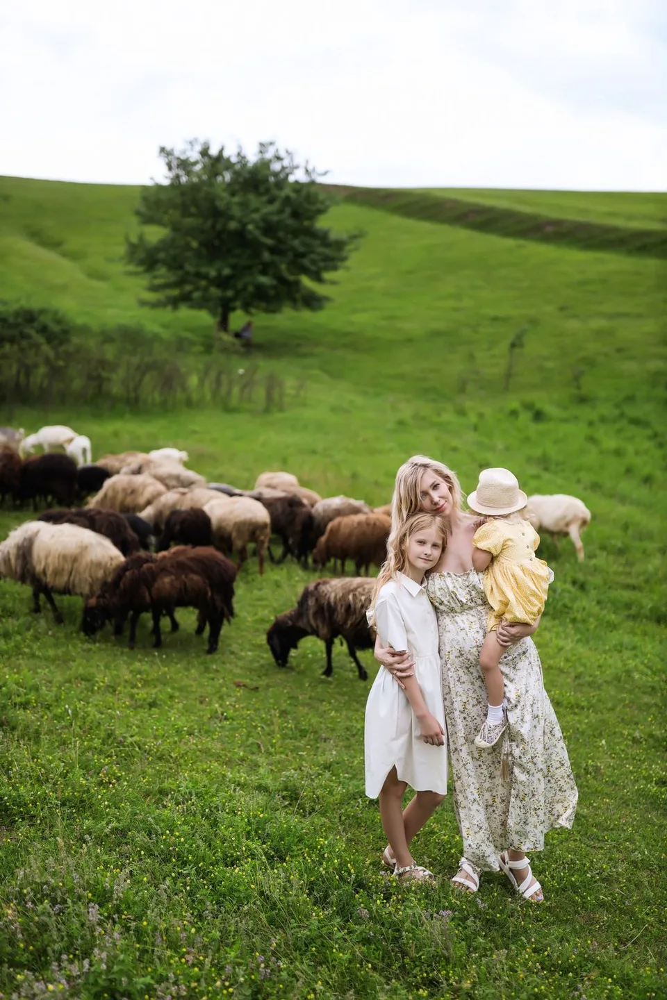 Children in light dresses stand in green meadow with sheep grazing in background.