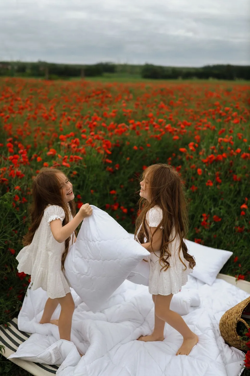Two people in white dresses having pillow fight on blanket in red poppy field.
