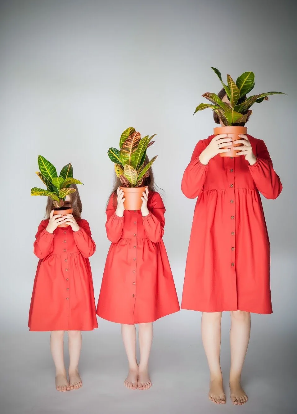 Three figures in matching coral dresses holding houseplants in front of their faces in ascending height order.