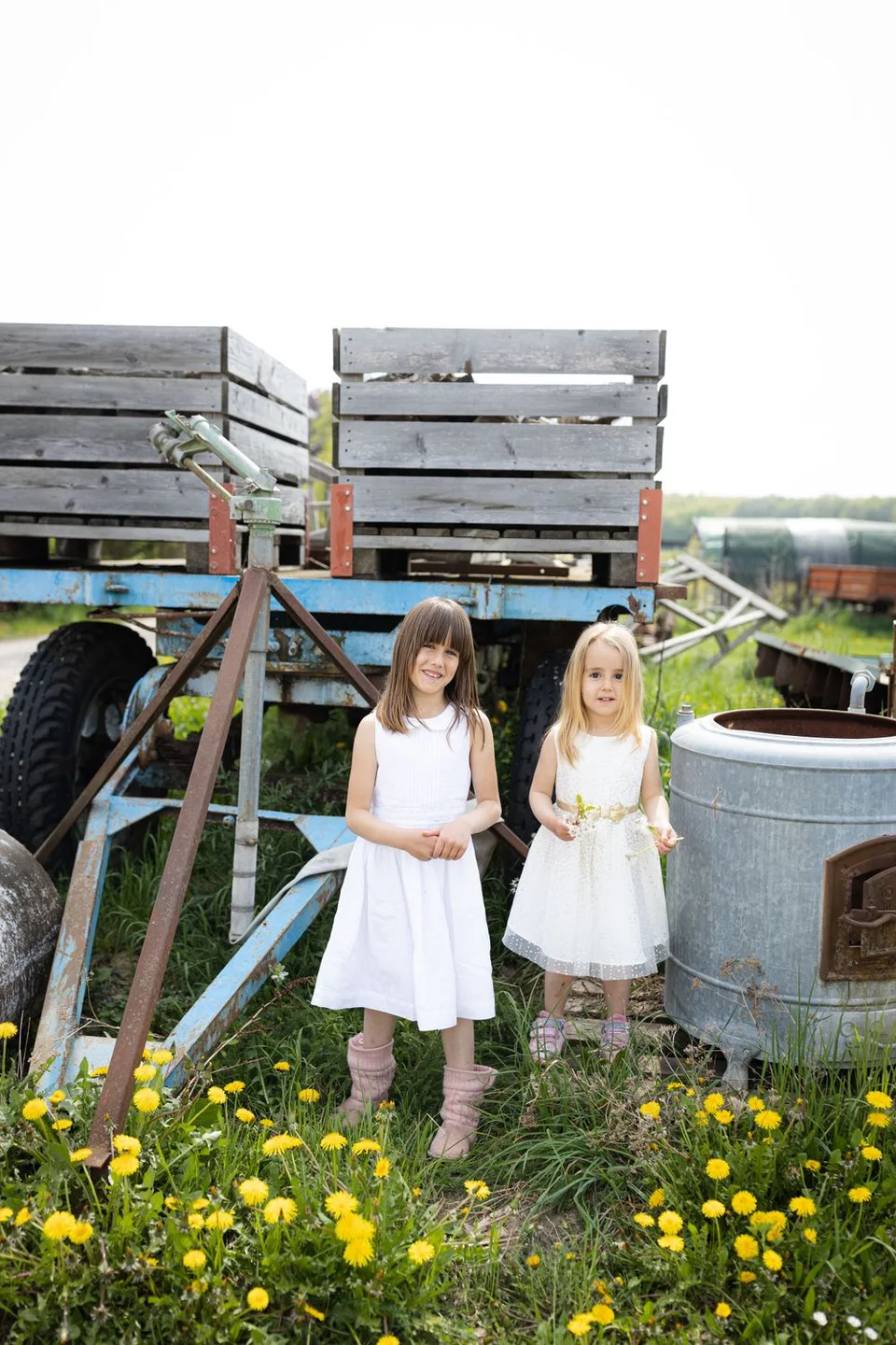 Two young people in white dresses stand by rustic wooden crates and yellow wildflowers.