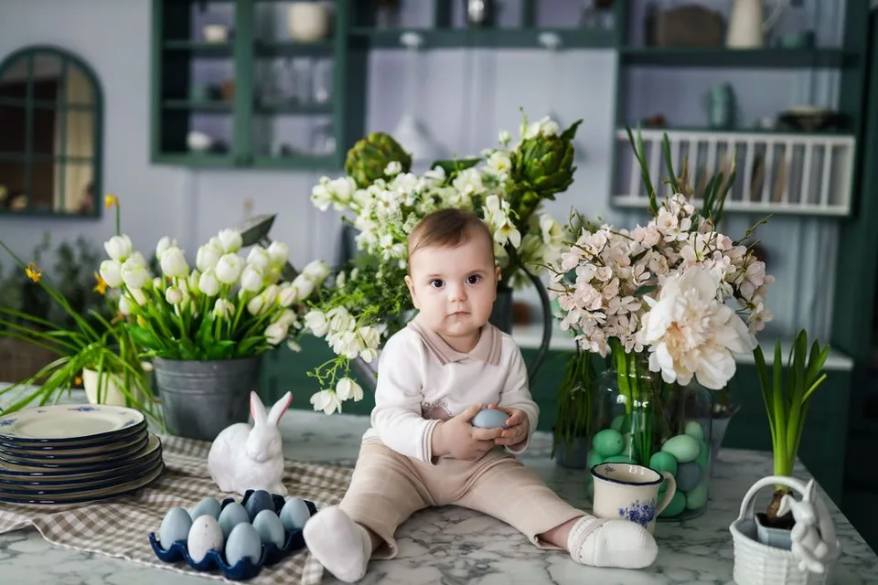 Baby sits among spring flowers and Easter decorations on rustic table with white bunny and blue eggs.