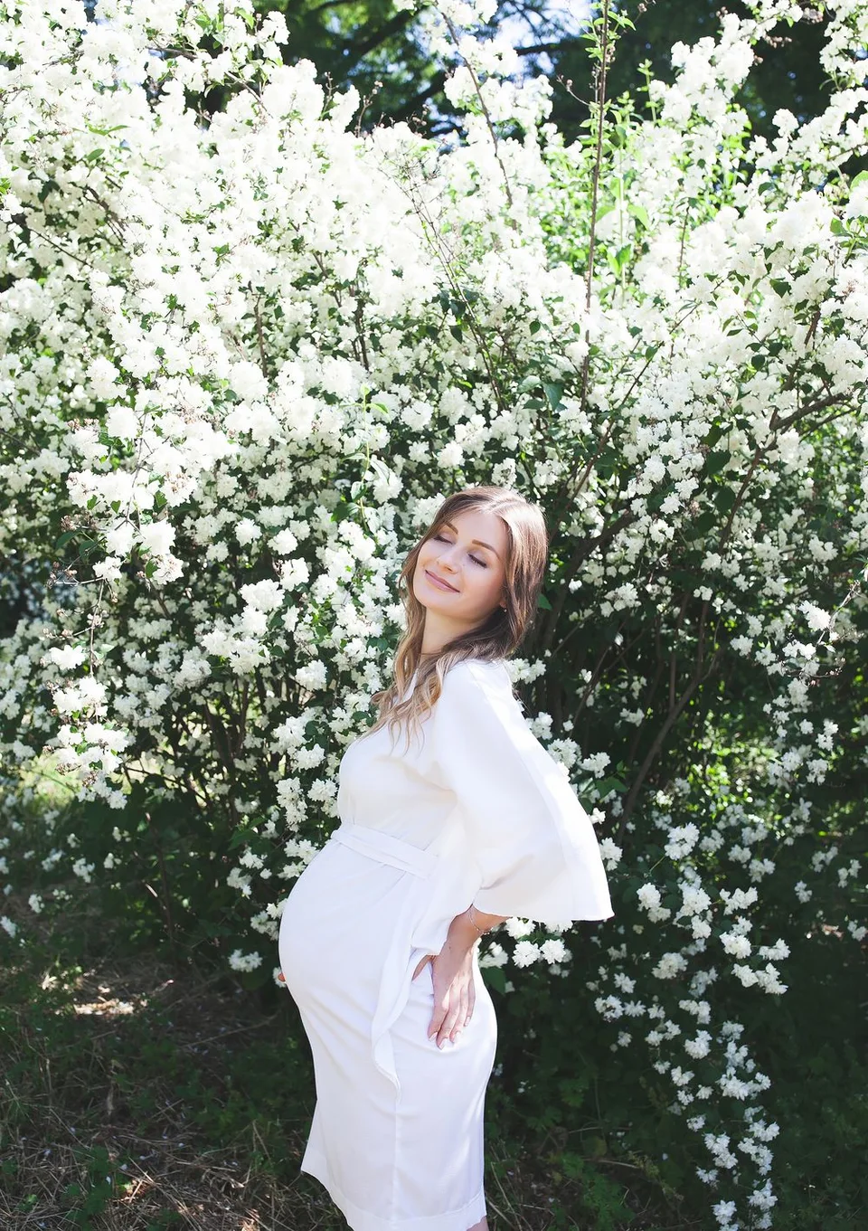 A dreamy maternity portrait in a white dress against blooming white flowers in a garden setting.