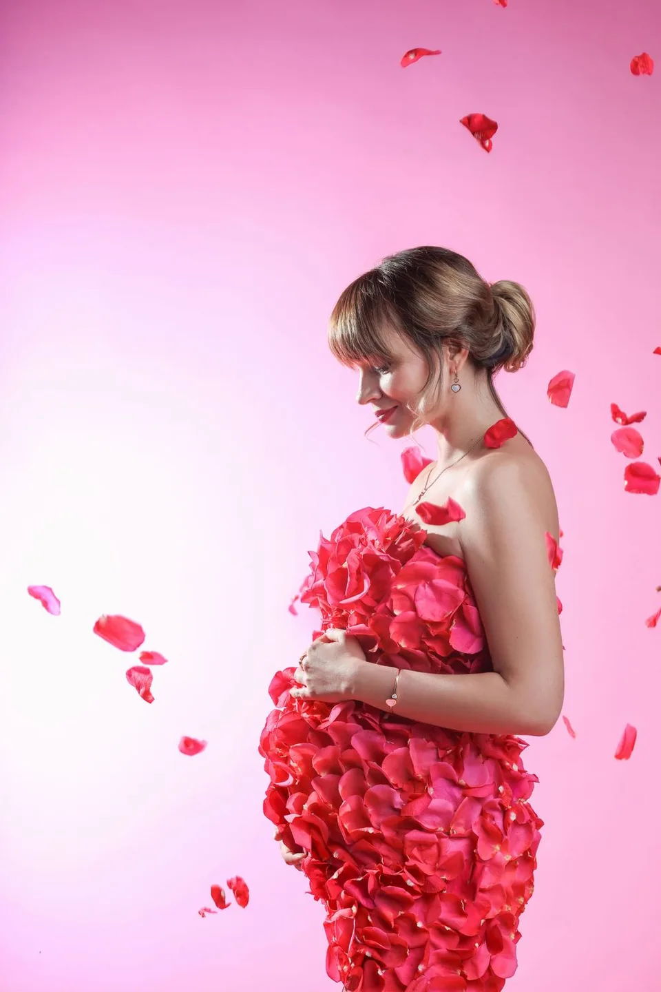 Woman in dress made of pink rose petals with falling petals against pink background.