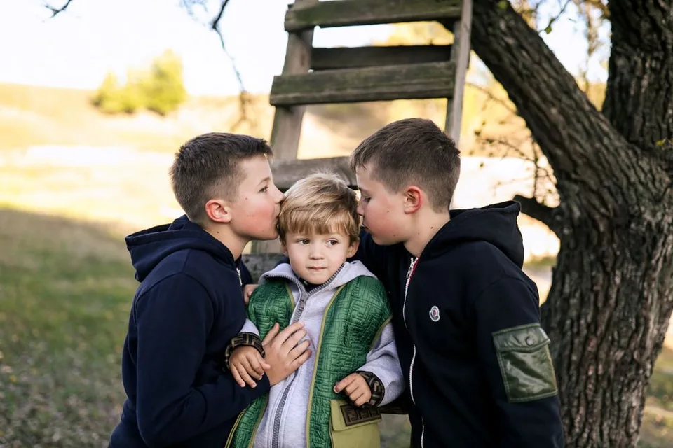 Three children in navy hoodies sharing secrets outdoors near wooden ladder.