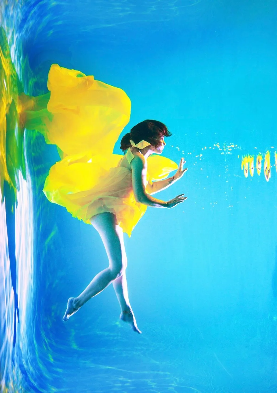 Artistic underwater photo of a dancer in a flowing yellow dress against bright blue background.