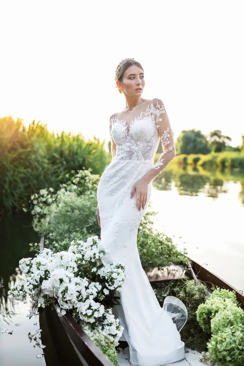 Fitted lace wedding gown with floral decorations photographed in wooden boat on tranquil water.