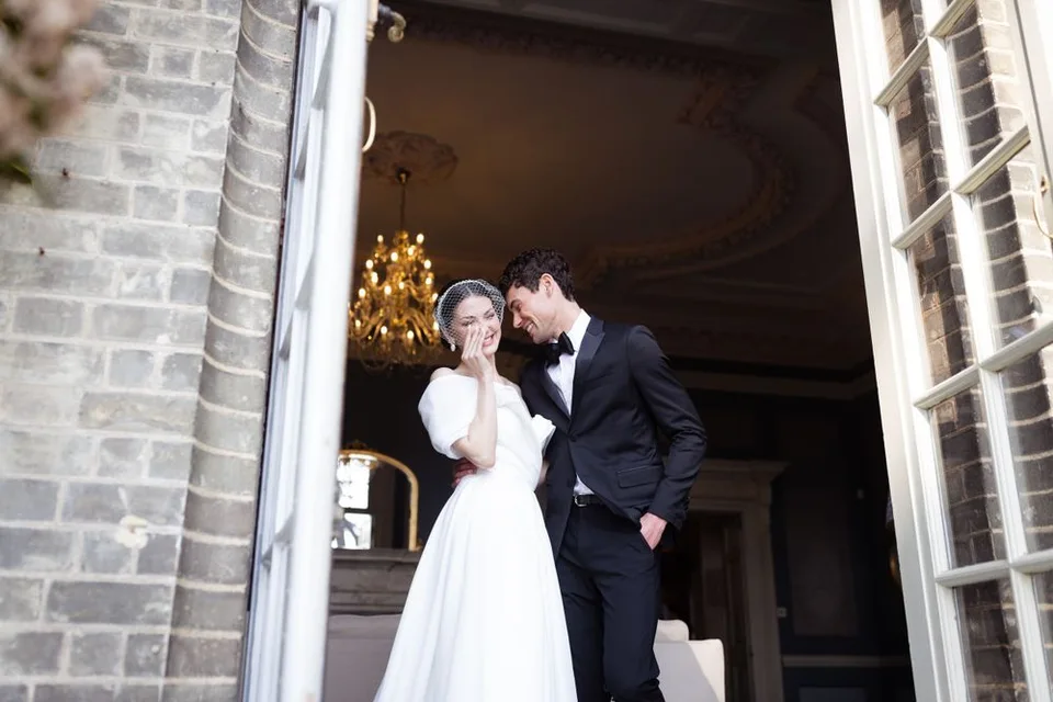 Newlyweds share an intimate moment framed by stone doorway in elegant wedding attire.