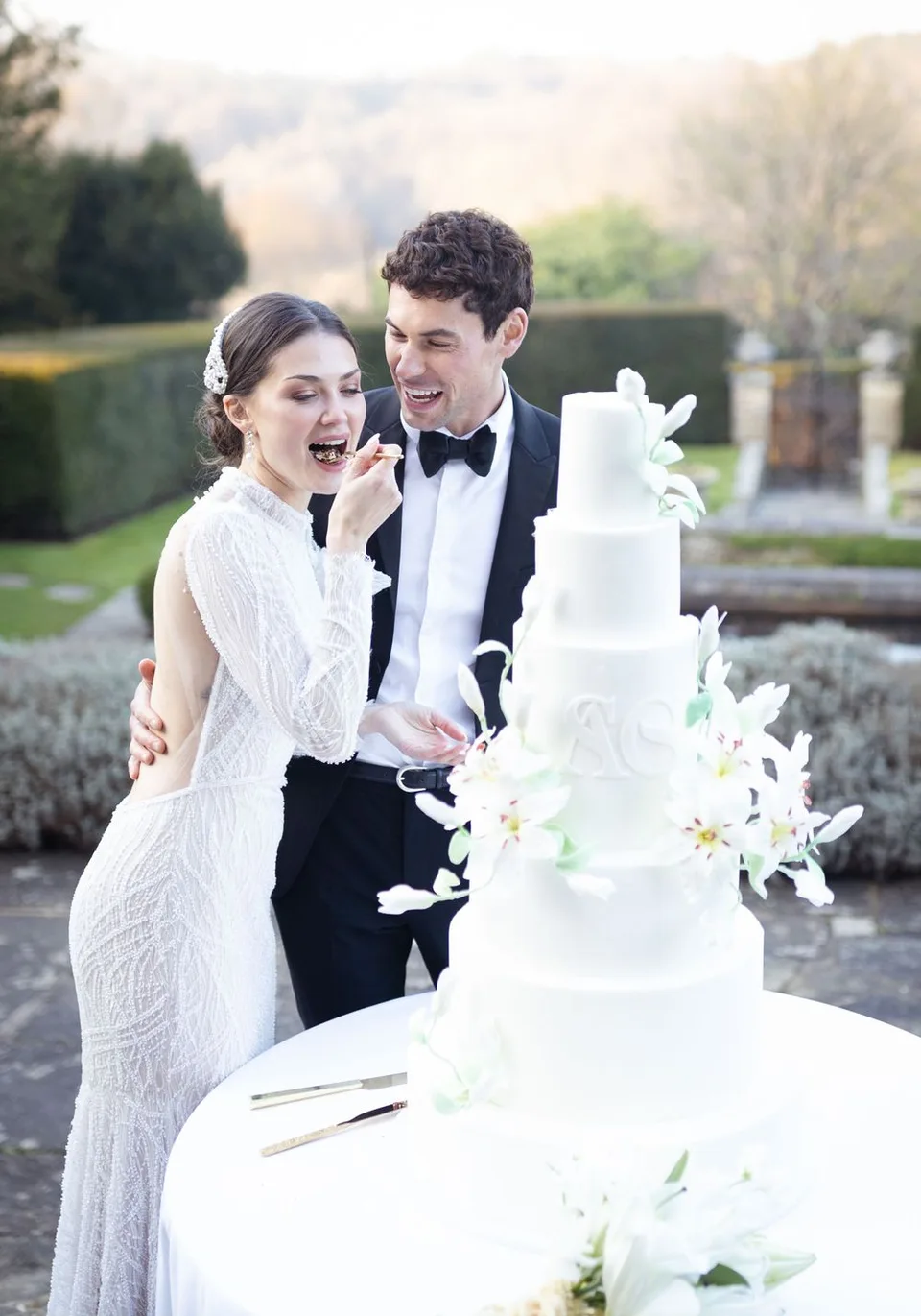 Elegant white tiered wedding cake decorated with sugar flowers being enjoyed at an outdoor reception.