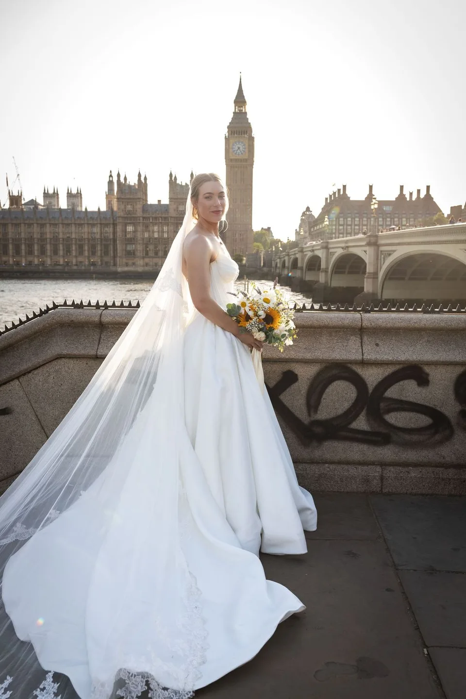 Bride in white gown with veil holds bouquet by Westminster Bridge with Big Ben and Houses of Parliament, London.