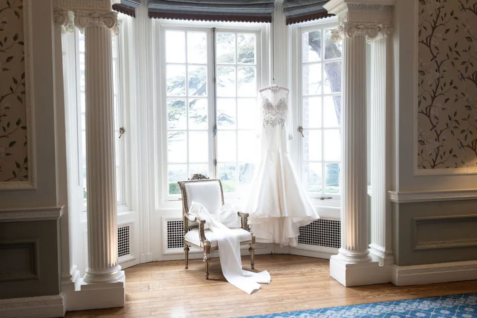 Elegant bridal gown displayed in a sunlit bay window room with classical columns and hardwood floors.