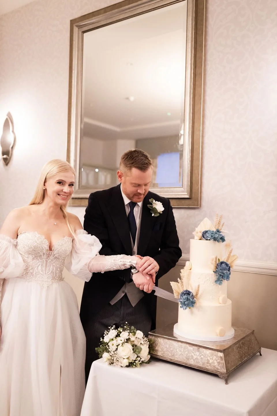 Bride and groom cutting a three-tier white wedding cake with blue floral decorations at their elegant wedding reception.
