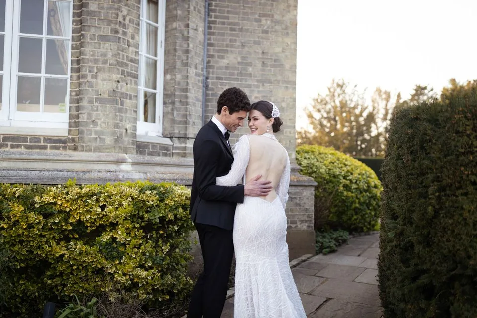 A couple embraces outside a brick mansion surrounded by manicured hedges during their wedding photoshoot.