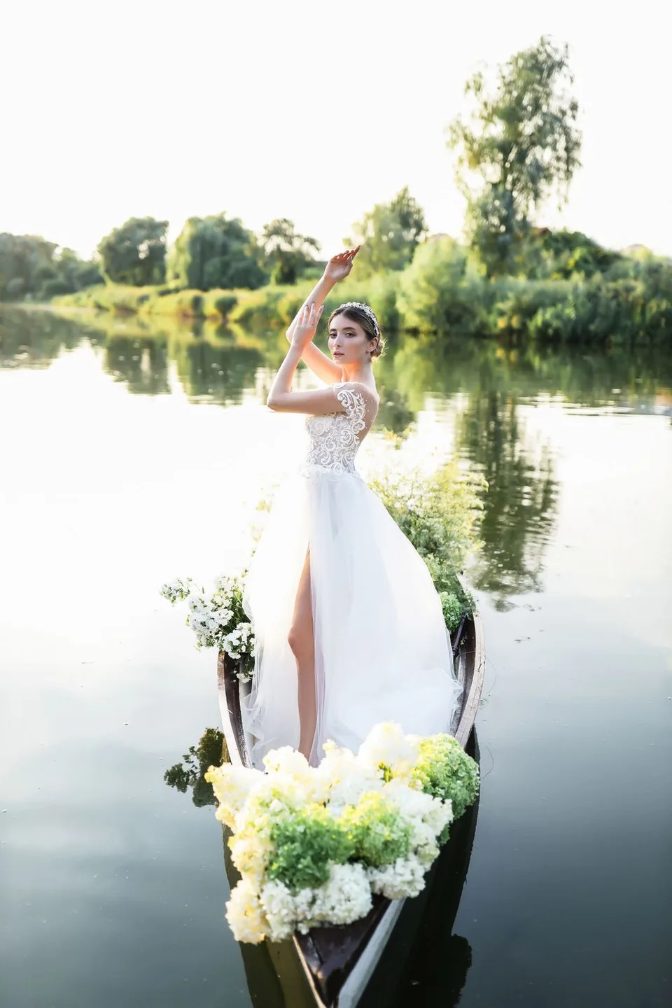 White lace wedding dress photographed in a flower-decorated boat on a serene lake at sunset.