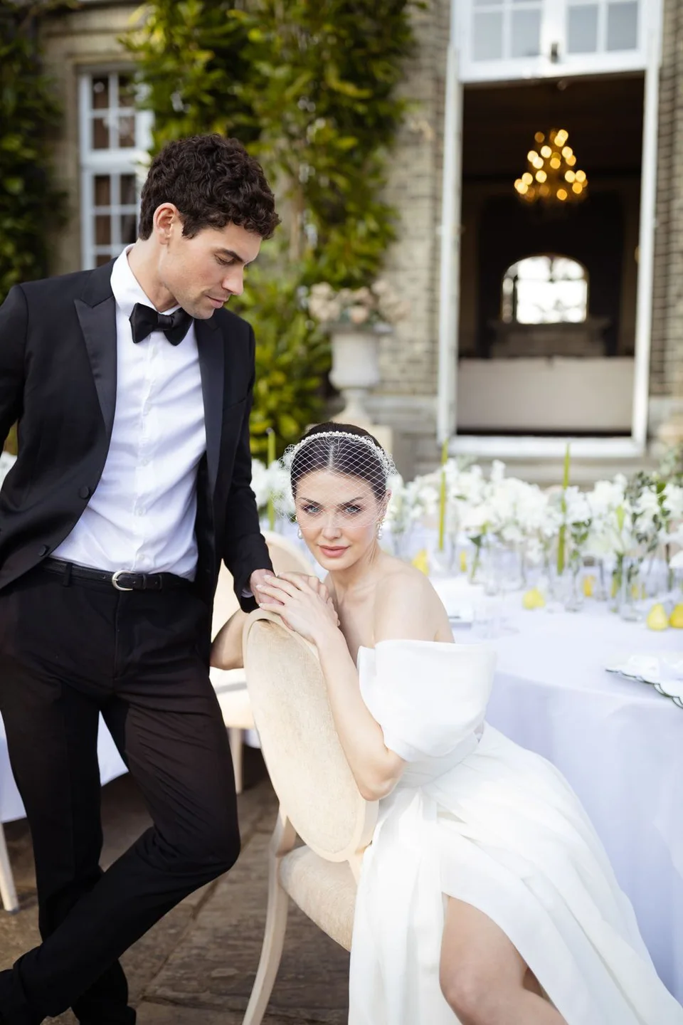 Wedding couple poses at outdoor table setting with chandelier visible through doorway.