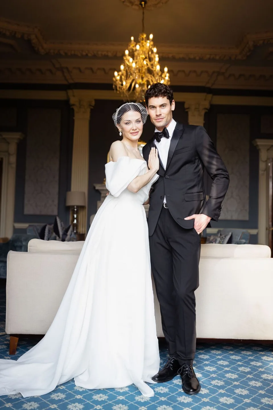 Elegant wedding portrait with bride in white gown and groom in tuxedo under crystal chandelier.