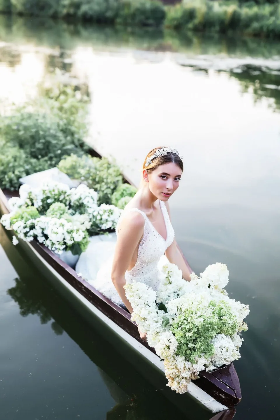 Bridal portrait in vintage boat filled with white floral arrangements on calm water.