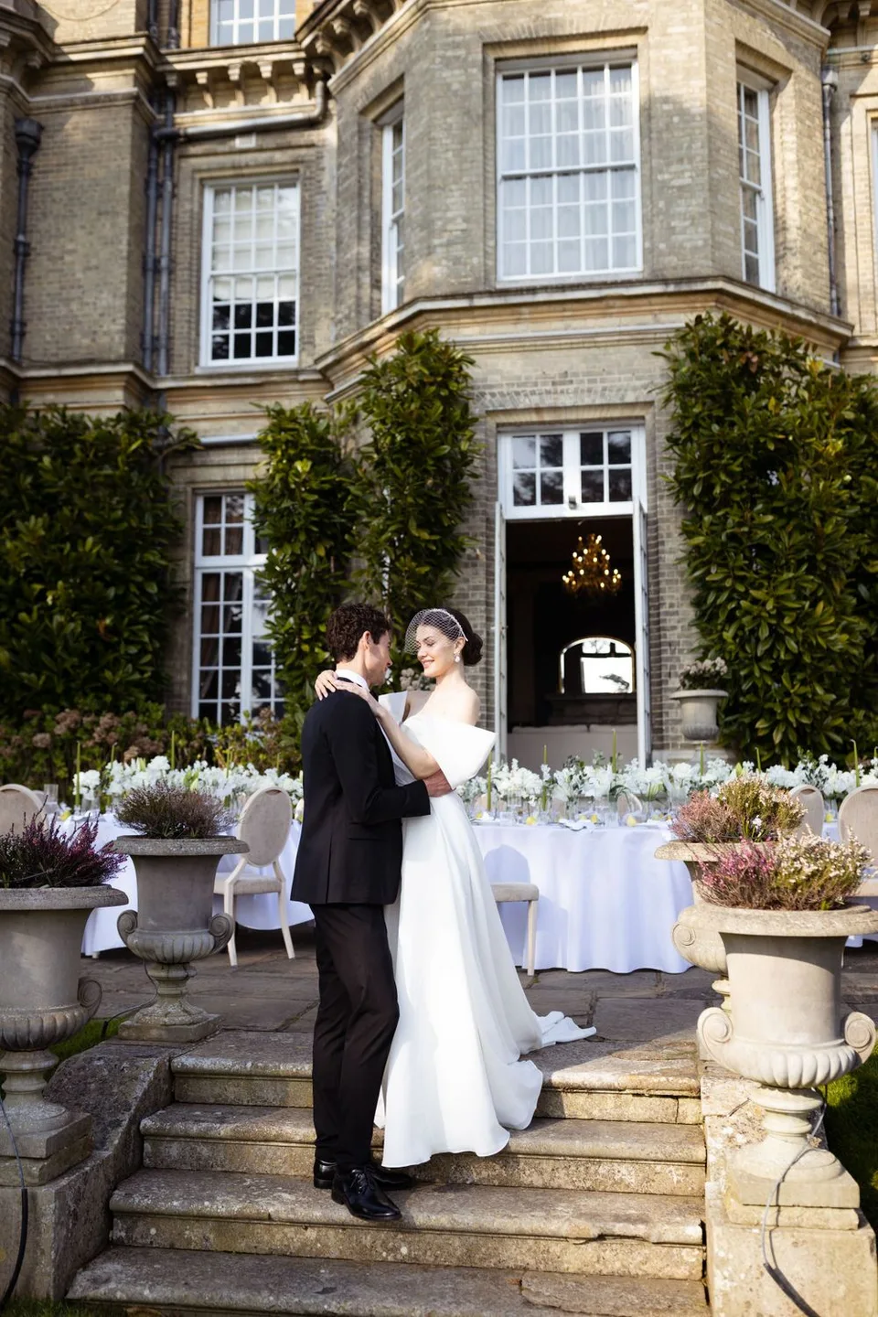 A wedding couple stands on stone steps outside an elegant manor house with tables set for an outdoor reception.