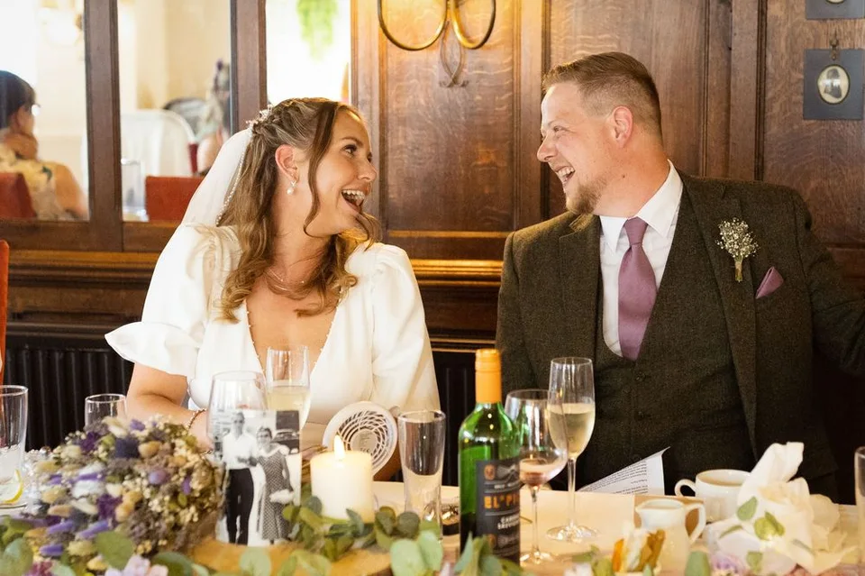 A happy couple shares a joyful moment together at their wedding reception table decorated with flowers and wine glasses.