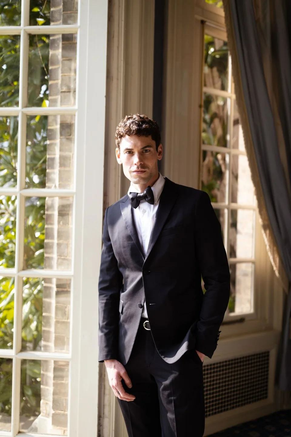 Person in black tuxedo and bow tie posing by elegant window with natural light.