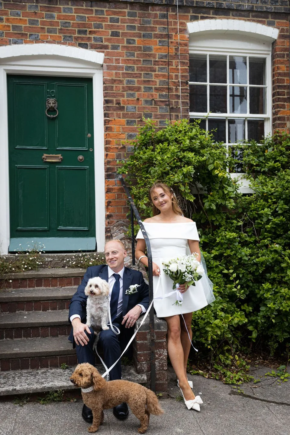 A couple poses outside a brick house with their small dogs in a series of casual photos by a green front door.
