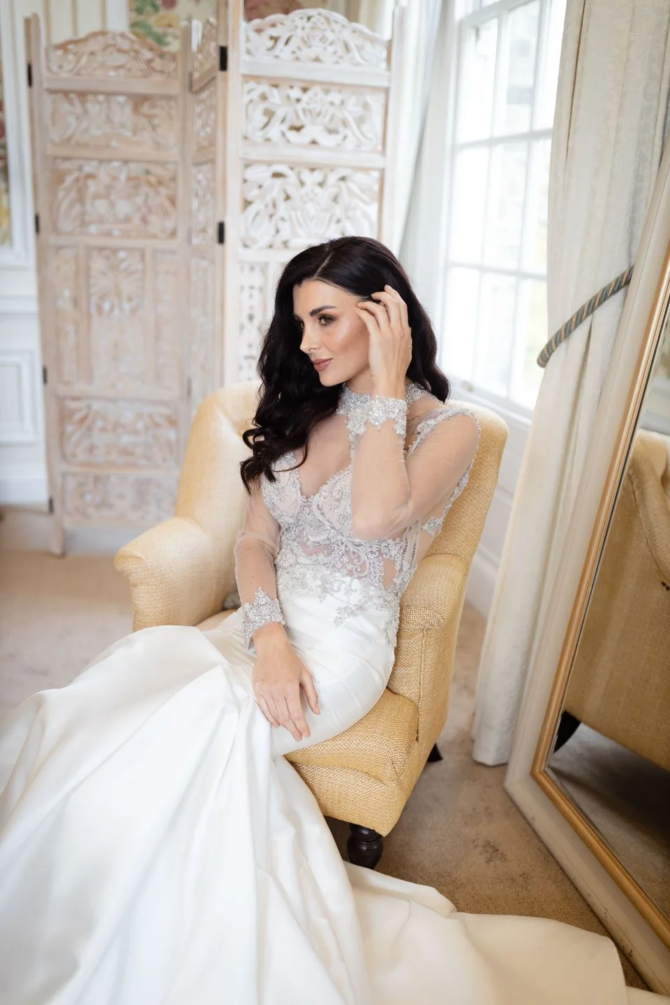 A bride in an intricate lace gown poses elegantly on a yellow chair in front of decorative room dividers.