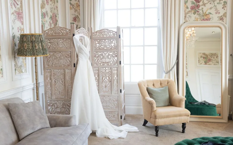 Elegant bridal gown displayed in a luxurious dressing room with ornate screen and full-length mirror.
