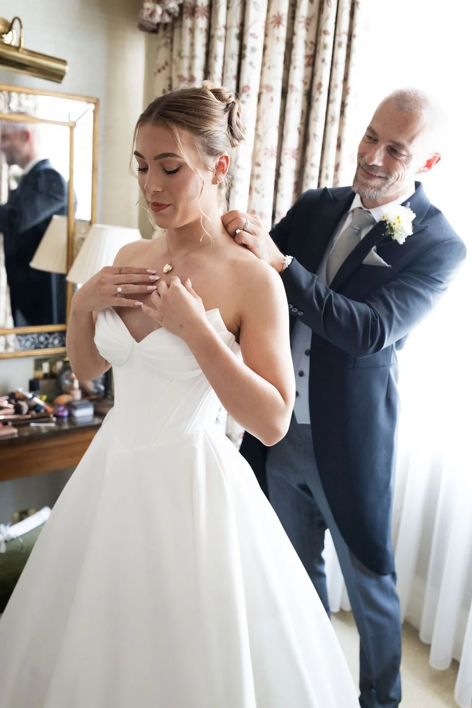 Father of the bride helping daughter with necklace in bridal suite before wedding ceremony.