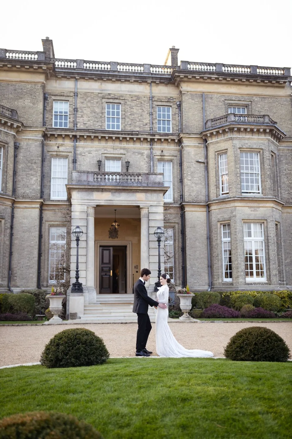 Wedding couple stands before grand stone mansion entrance with manicured topiary gardens.