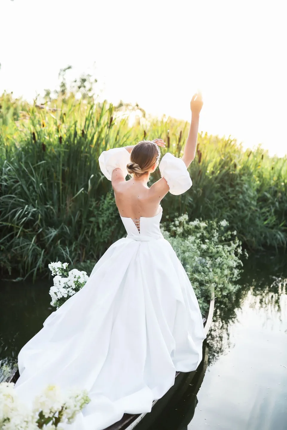 Bride in flowing white ballgown raises her arms triumphantly by a sunlit pond.