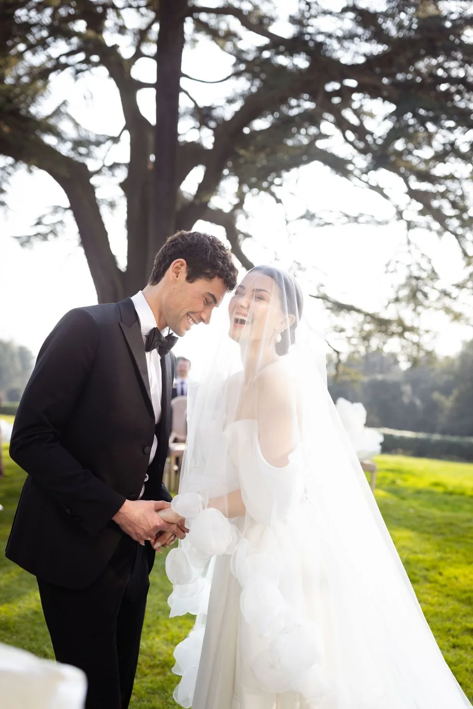 A joyful wedding moment between newlyweds under a large tree in afternoon sunlight.