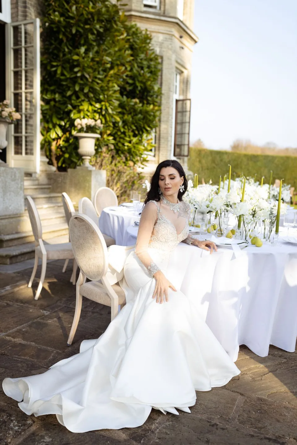 Bride in flowing white gown sits at luxurious outdoor table setting with mansion in background.
