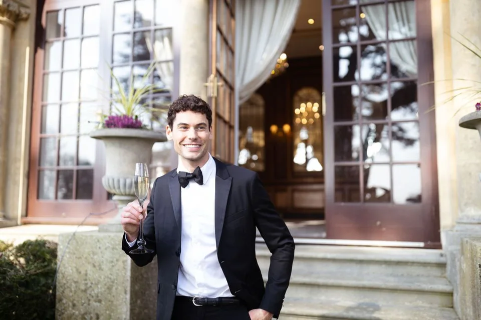 Dapper groom in classic tuxedo and bowtie poses with champagne glass outside luxury venue.