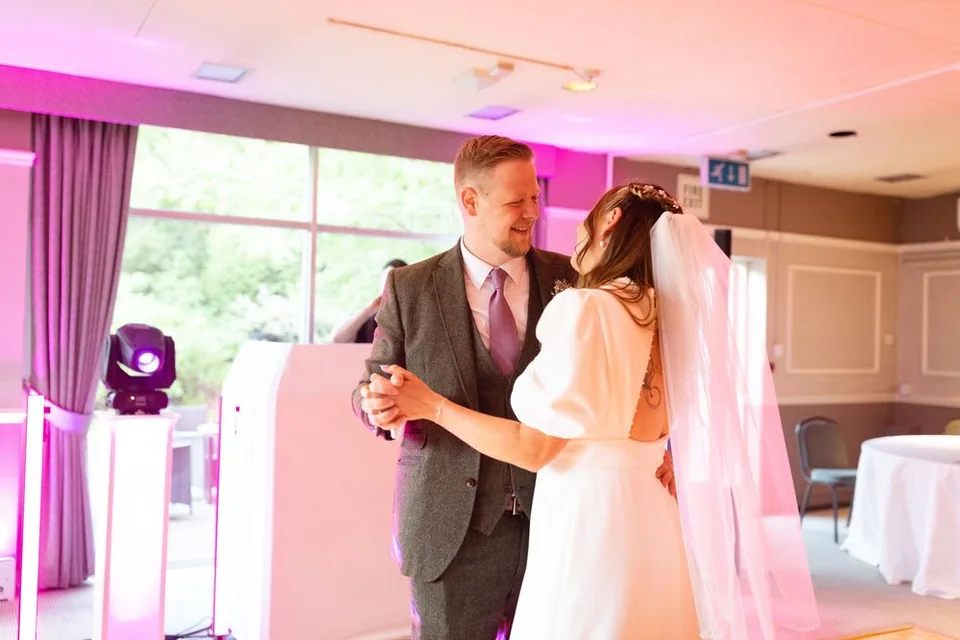 A couple dances in a formal setting with pink-tinted lighting, the bride wearing a white dress and flowing veil.