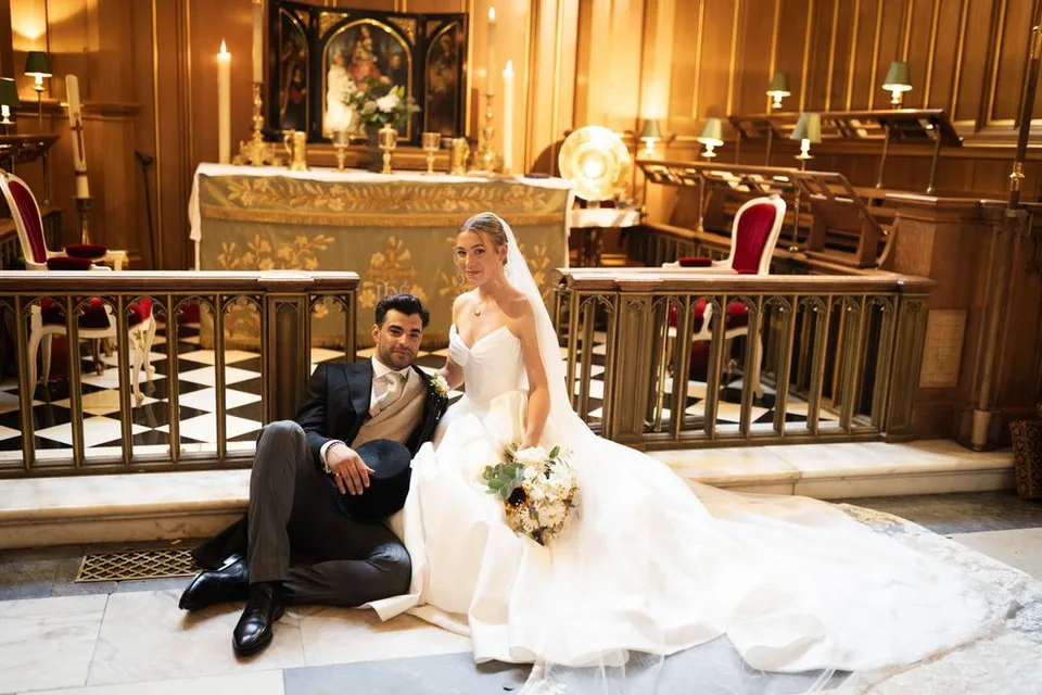 Bride and groom posing together on church altar steps, bride in ballgown holding bouquet, groom in dark suit.