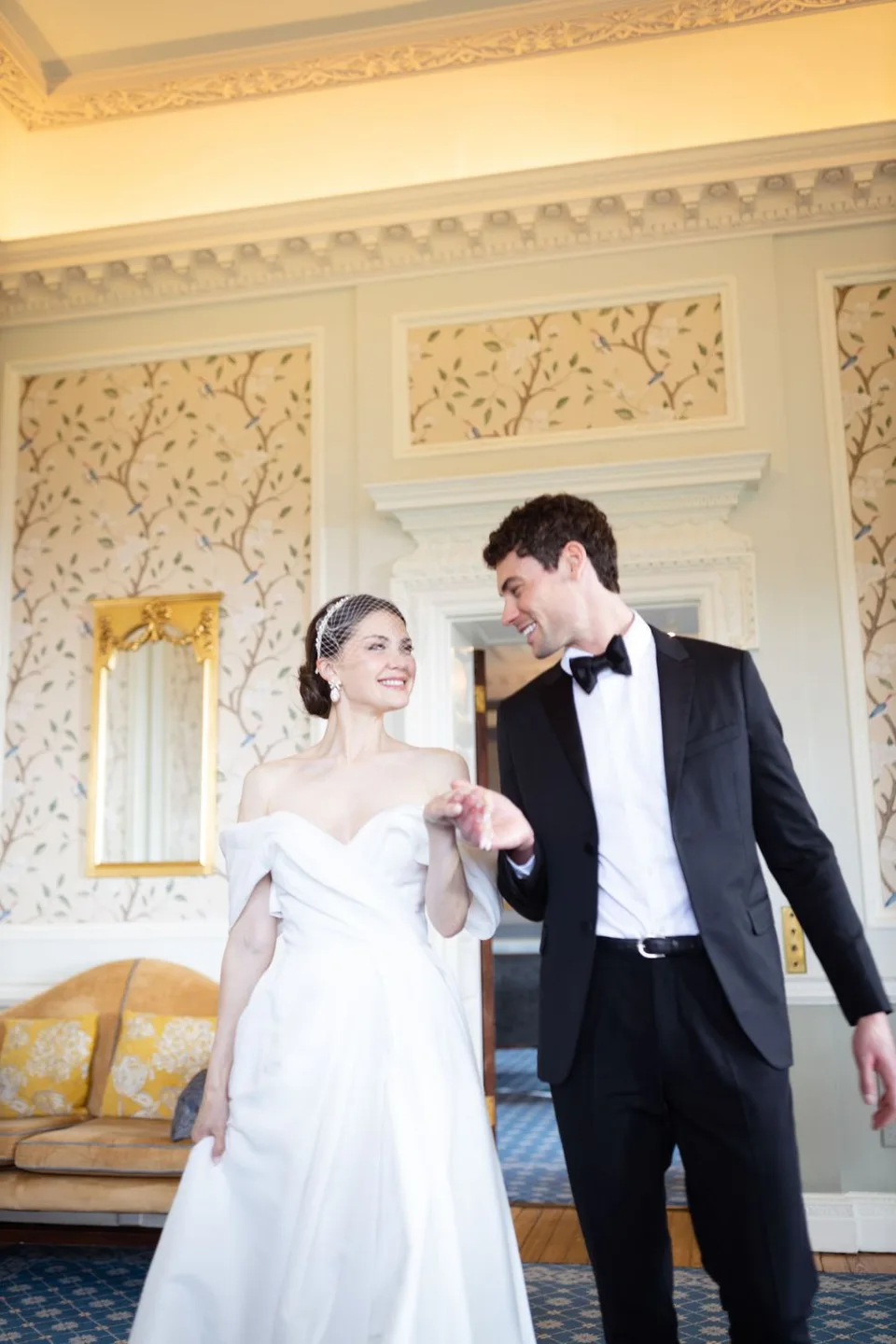 Elegant couple dances in ornate room with floral wallpaper and gold accents.