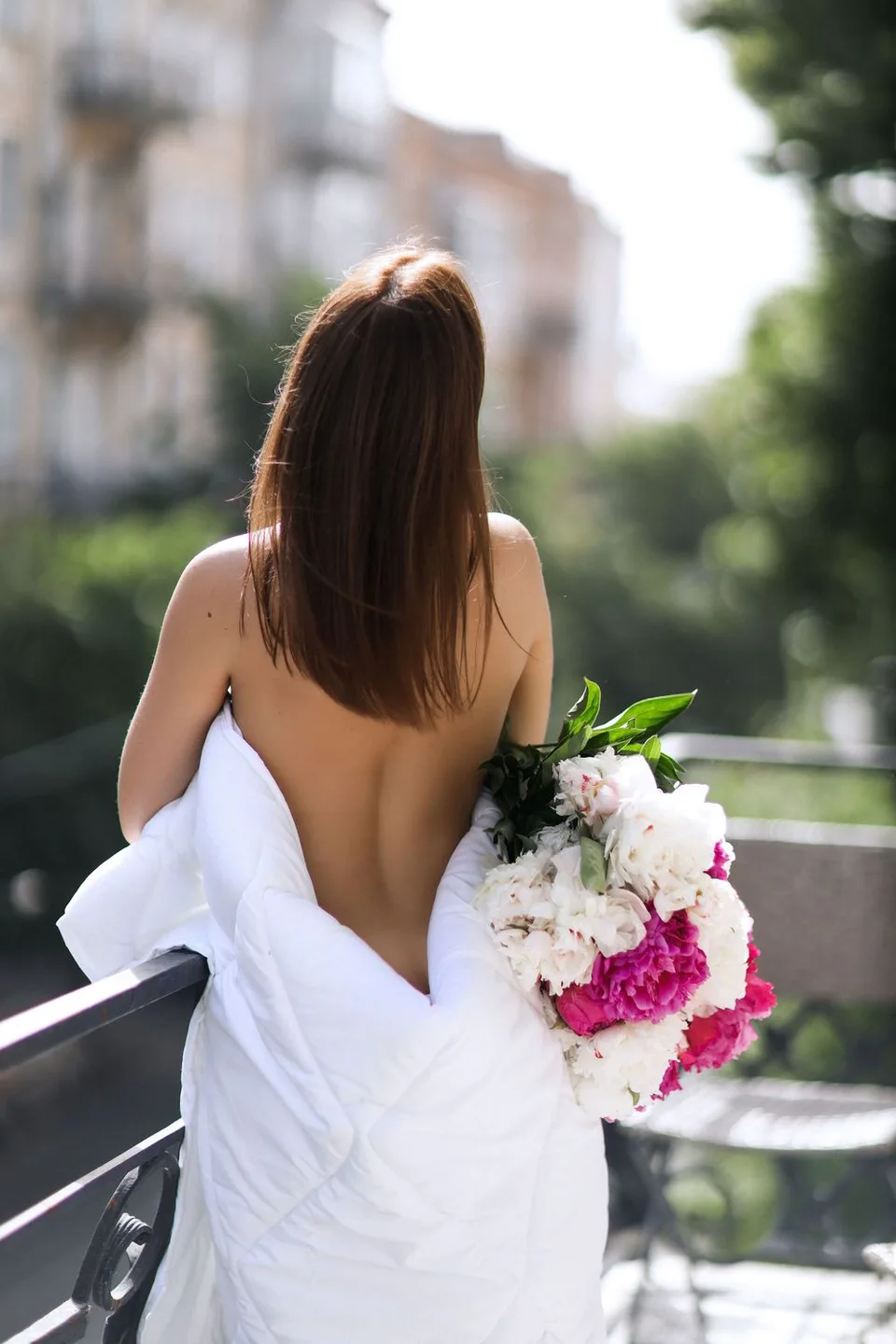 Back view of person in white dress holding pink and white bouquet on balcony.
